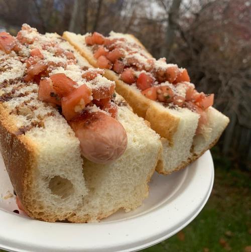 Two hot dogs topped with diced tomatoes and cheese, served on a white plate against a blurred outdoor background.
