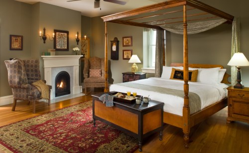 Cozy bedroom with a four-poster bed, fireplace, armchairs, and warm lighting, featuring elegant decor and a rug.