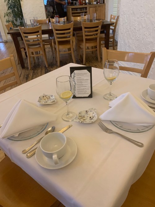 A neatly set dining table with white tablecloth, plates, cutlery, and a menu, in a cozy restaurant setting.