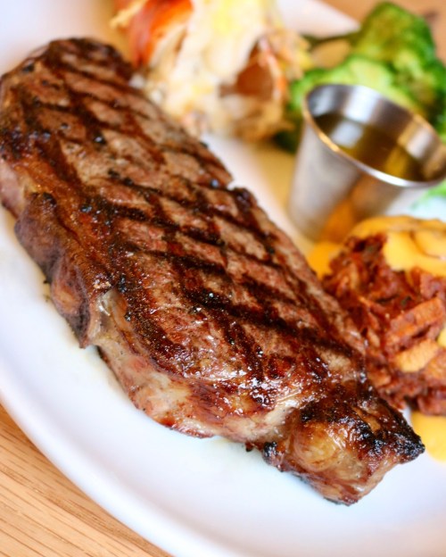 A grilled steak with a seared crust, served with broccoli and a side of shredded potatoes on a white plate.