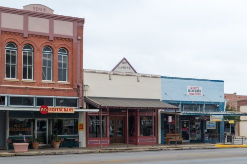 Historic storefronts line a street, featuring a restaurant and a market, with a cloudy sky overhead.