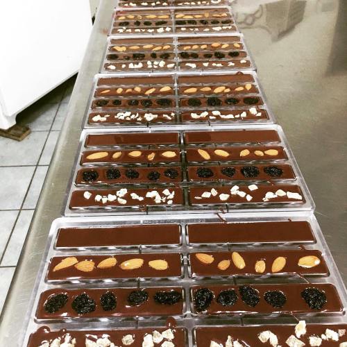 Rows of chocolate bars in molds, topped with almonds, dried fruits, and oats, on a stainless steel surface.