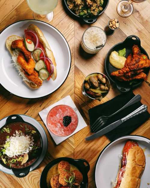 A spread of various dishes including a hot dog, fried chicken, cocktails, and sides on a wooden table.