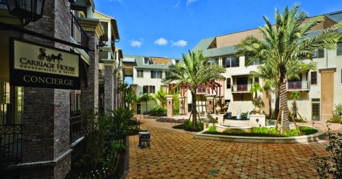 A sunny courtyard with palm trees, a fountain, and a sign for Carriage House Condominiums & Suites.