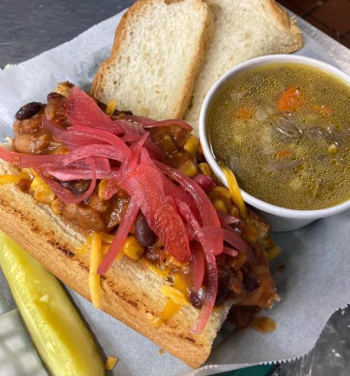 A hearty sandwich topped with chili, cheese, and pickled onions, served with a side of soup and a pickle.