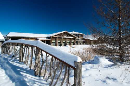 A snow-covered landscape featuring a wooden bridge leading to a modern building against a clear blue sky.