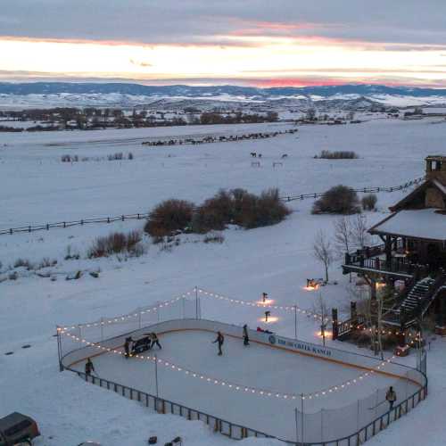 A snowy landscape with an outdoor ice rink, surrounded by trees and mountains, at sunset with lights strung around.