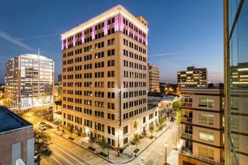 A historic building illuminated in pink lights at dusk, surrounded by modern city architecture and streets.