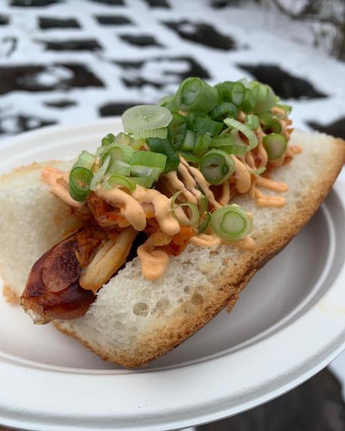 A close-up of a hot dog in a bun, topped with green onions and a drizzle of sauce, served on a paper plate.