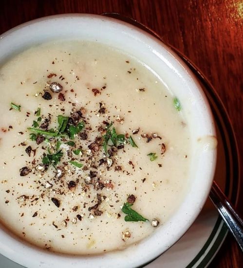 A bowl of creamy soup topped with herbs and black pepper, placed on a wooden table.
