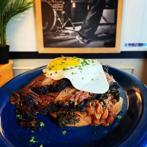 A plate featuring a slice of bread topped with brisket, beans, greens, and a fried egg, with a blurred background of a musician.