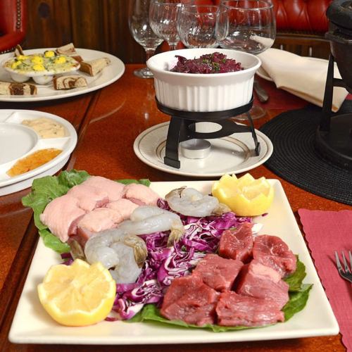 A platter of raw meats and shrimp on lettuce, with lemon slices, alongside a fondue pot and other dishes in the background.