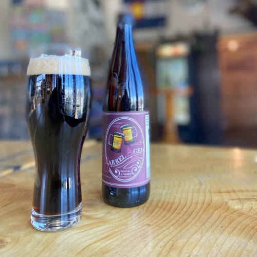 A glass of dark beer with a frothy head next to a bottle labeled "Barrel Aged" on a wooden table.