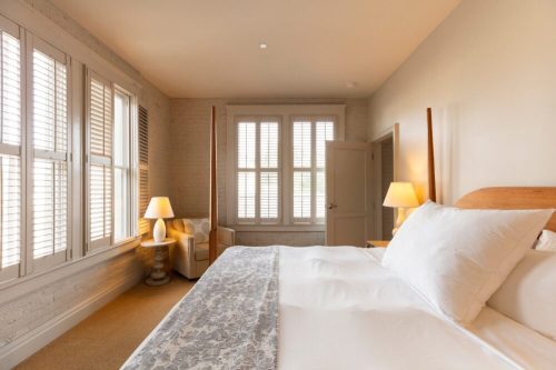 Bright, cozy bedroom with a large bed, two lamps, and windows with white shutters. Soft, neutral color palette.