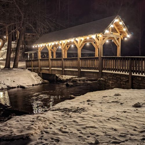 A wooden bridge with lights spans a calm stream, surrounded by trees at night. Snow covers the ground.