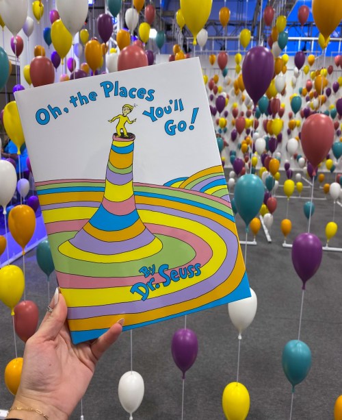 A person holds the book "Oh, the Places You'll Go!" by Dr. Seuss, surrounded by colorful balloons in a festive setting.