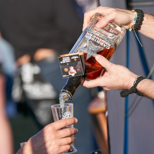 A person pours whiskey from a bottle into a shot glass held by another person at an outdoor event.