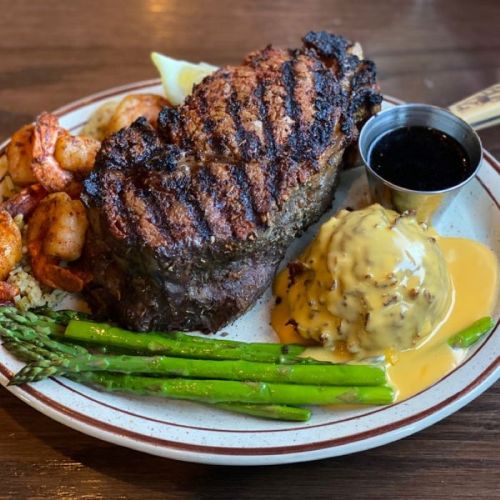 A plate featuring a grilled steak, shrimp, asparagus, mashed potatoes with cheese, and a small cup of sauce.
