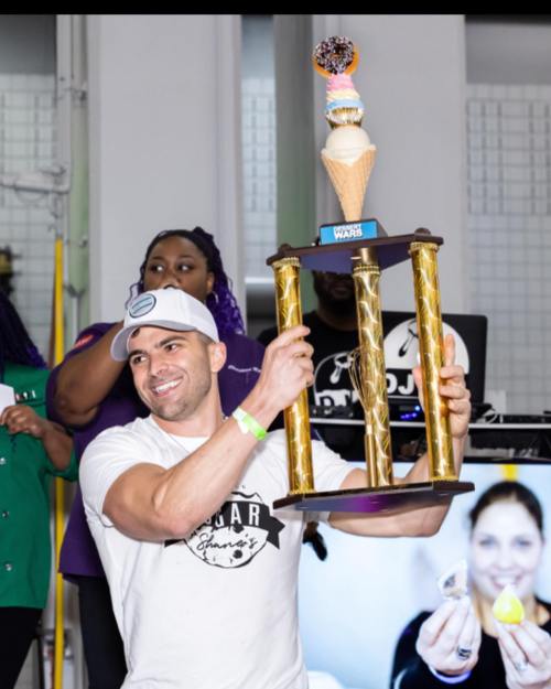 A man in a white shirt holds a large ice cream trophy, smiling, with a crowd and a DJ booth in the background.