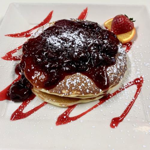 Stack of pancakes topped with berry sauce, powdered sugar, and garnished with a strawberry and orange slice.