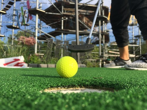 A close-up of a bright yellow golf ball on a green putting surface, with a person preparing to putt in the background.