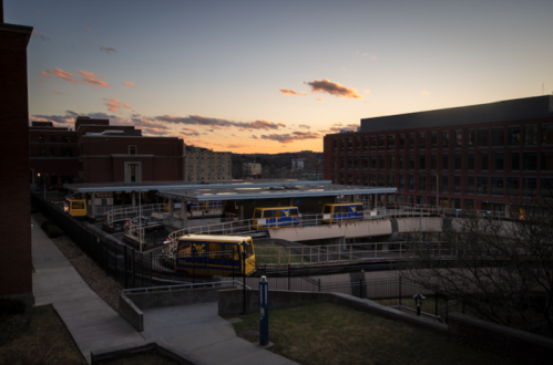 Sunset view of a university campus with buildings and a bus stop, featuring buses and a clear sky with scattered clouds.