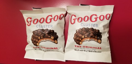 Two packages of GooGoo Cluster candy, featuring a chocolate-covered treat with a bite taken out, on a red background.