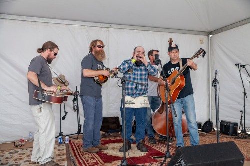 A group of five musicians perform together under a tent, playing various instruments and singing.