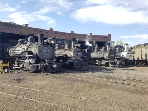 Three vintage steam locomotives parked in a roundhouse, with smoke rising against a blue sky.