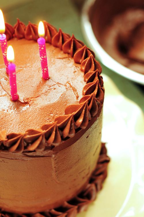 A chocolate cake with pink candles on top, decorated with swirls of frosting, sits on a plate.