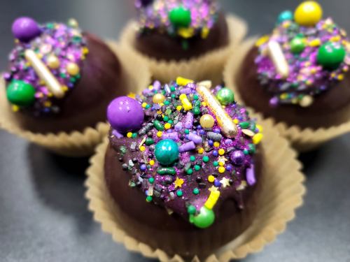 Colorful chocolate treats topped with vibrant sprinkles in purple, green, and gold, arranged in cupcake liners.