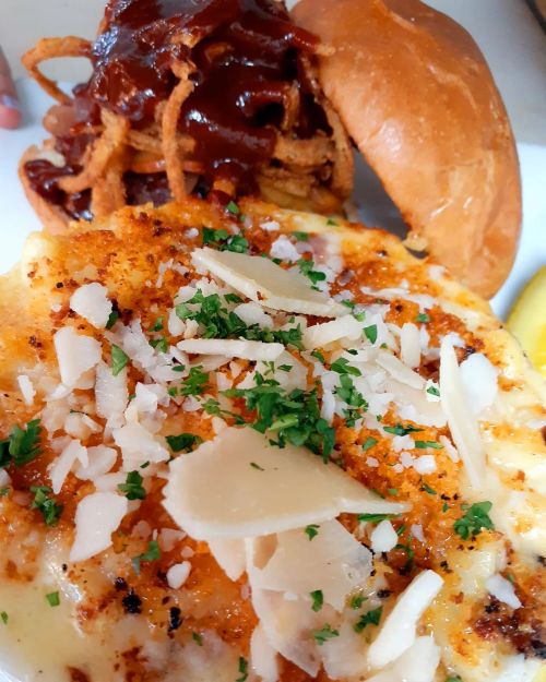 A close-up of a cheesy dish topped with herbs and cheese, alongside a burger with pulled pork and crispy onions.