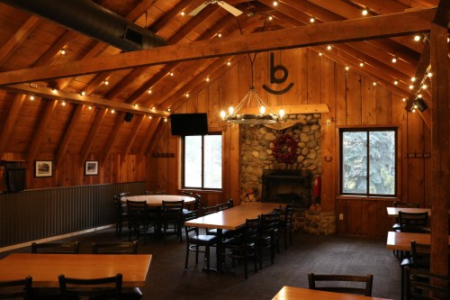 Cozy wooden interior of a dining hall with tables, a stone fireplace, and warm lighting.