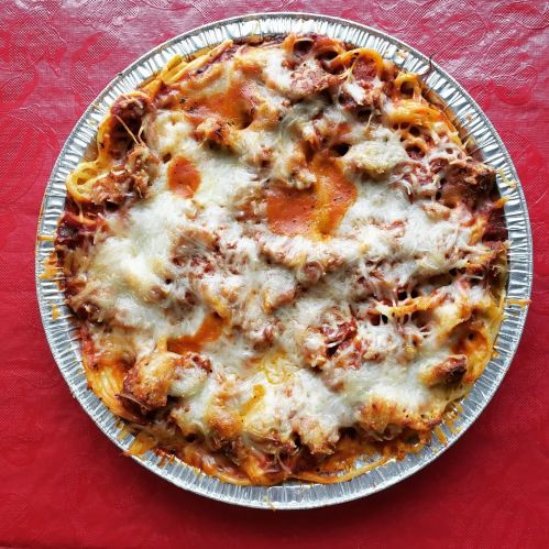 A baked pasta dish topped with melted cheese, served in a round aluminum tray on a red tablecloth.