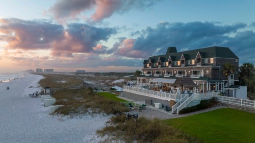 A beachfront hotel at sunset, with sandy shores, grassy areas, and a cloudy sky in the background.