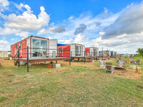 Colorful shipping container homes on stilts, surrounded by grass and chairs, under a blue sky with fluffy clouds.