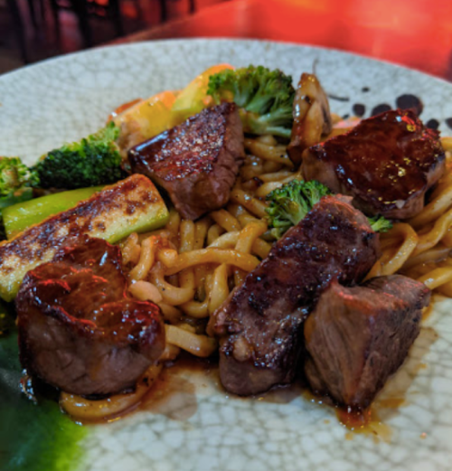 A plate of stir-fried noodles topped with tender beef chunks and colorful vegetables, garnished with a rich sauce.