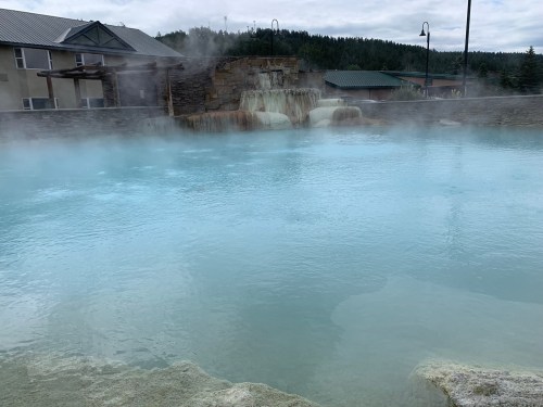A serene hot spring with steaming blue water, surrounded by rocks and a backdrop of trees and buildings.