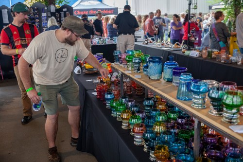 A man inspects colorful glassware at a bustling market, with many people browsing nearby stalls.