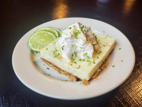 A slice of key lime pie topped with whipped cream and lime zest, served with lime slices on a white plate.