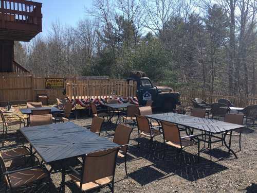 Outdoor seating area with tables and chairs, an old truck in the background, and a wooden fence surrounded by trees.