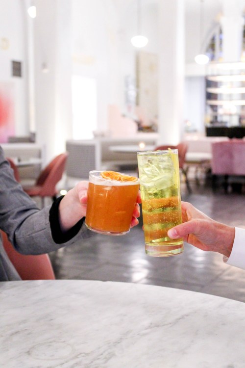 Two hands clink glasses in a stylish restaurant; one holds a cocktail, the other a sparkling drink.