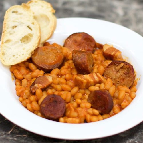 A bowl of baked beans with sliced sausage and pieces of meat, served with two slices of bread on the side.