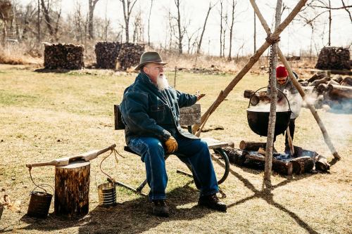 A man with a beard sits on a bench by a fire, watching a pot hanging over the flames, with stacks of wood in the background.