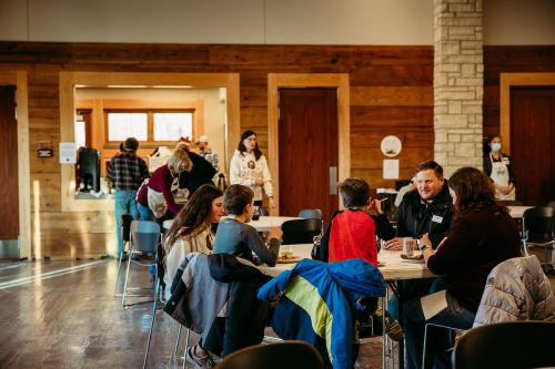 A group of people socializing at tables in a warm, wooden interior, with a kitchen area in the background.
