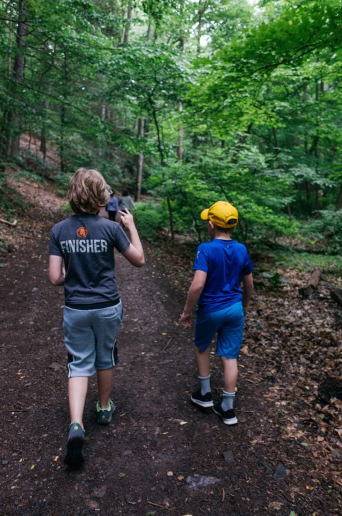 Two children walking on a wooded trail, surrounded by lush green trees. One wears a gray shirt and the other a yellow cap.