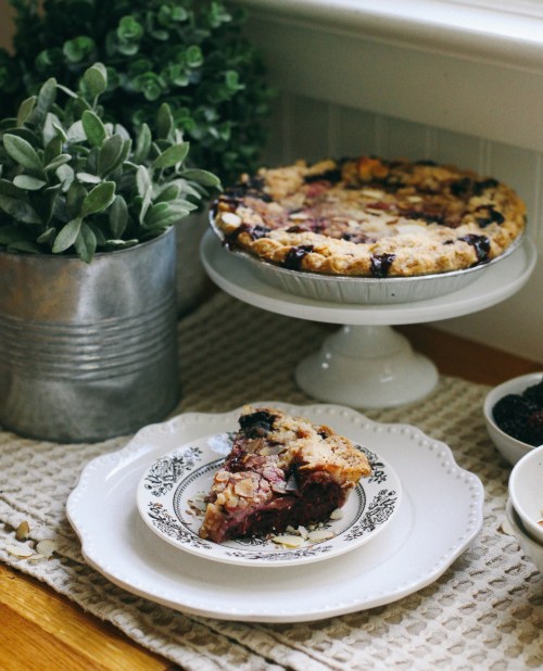 A slice of berry pie on a decorative plate, with the whole pie on a stand and a potted plant in the background.