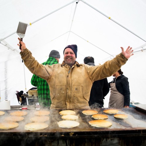 A smiling man in a tan jacket holds a spatula, standing in a tent with pancakes cooking on a griddle.