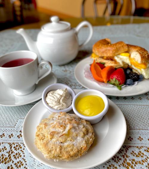 A cozy breakfast scene featuring a scone, lemon curd, whipped cream, fresh fruit, and a cup of tea.