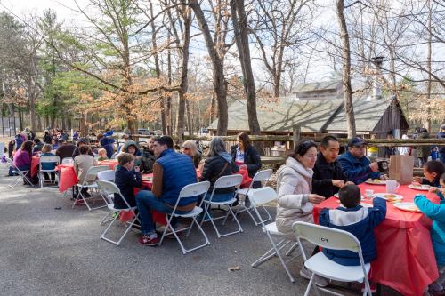 A group of people enjoying a meal outdoors at tables, surrounded by trees and a rustic building in the background.